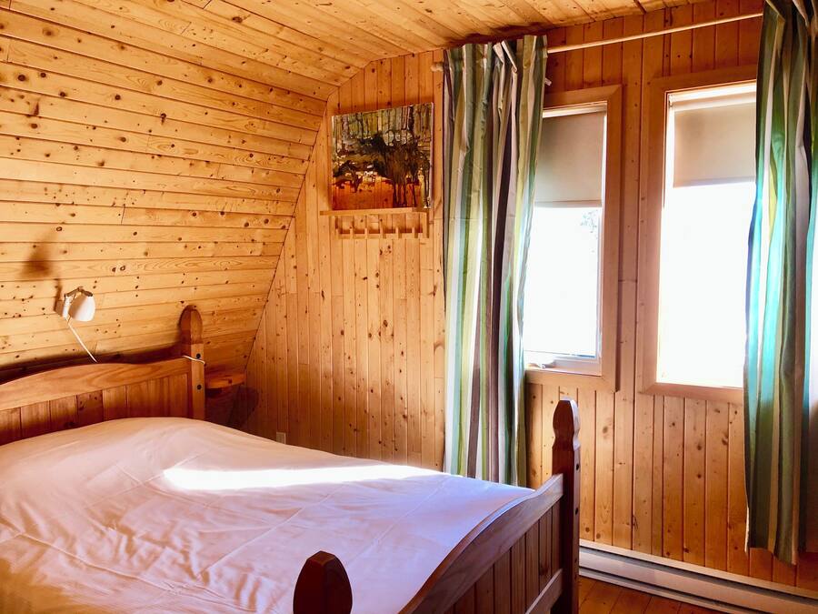 A wooden cabin bedroom with a bed, a window with curtains, and a wall-mounted light fixture.