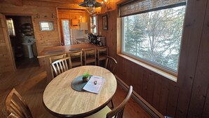 Interior acogedor de una cabaña con una mesa de comedor de madera, sillas, una zona de cocina y un amplio ventanal con vistas a árboles.