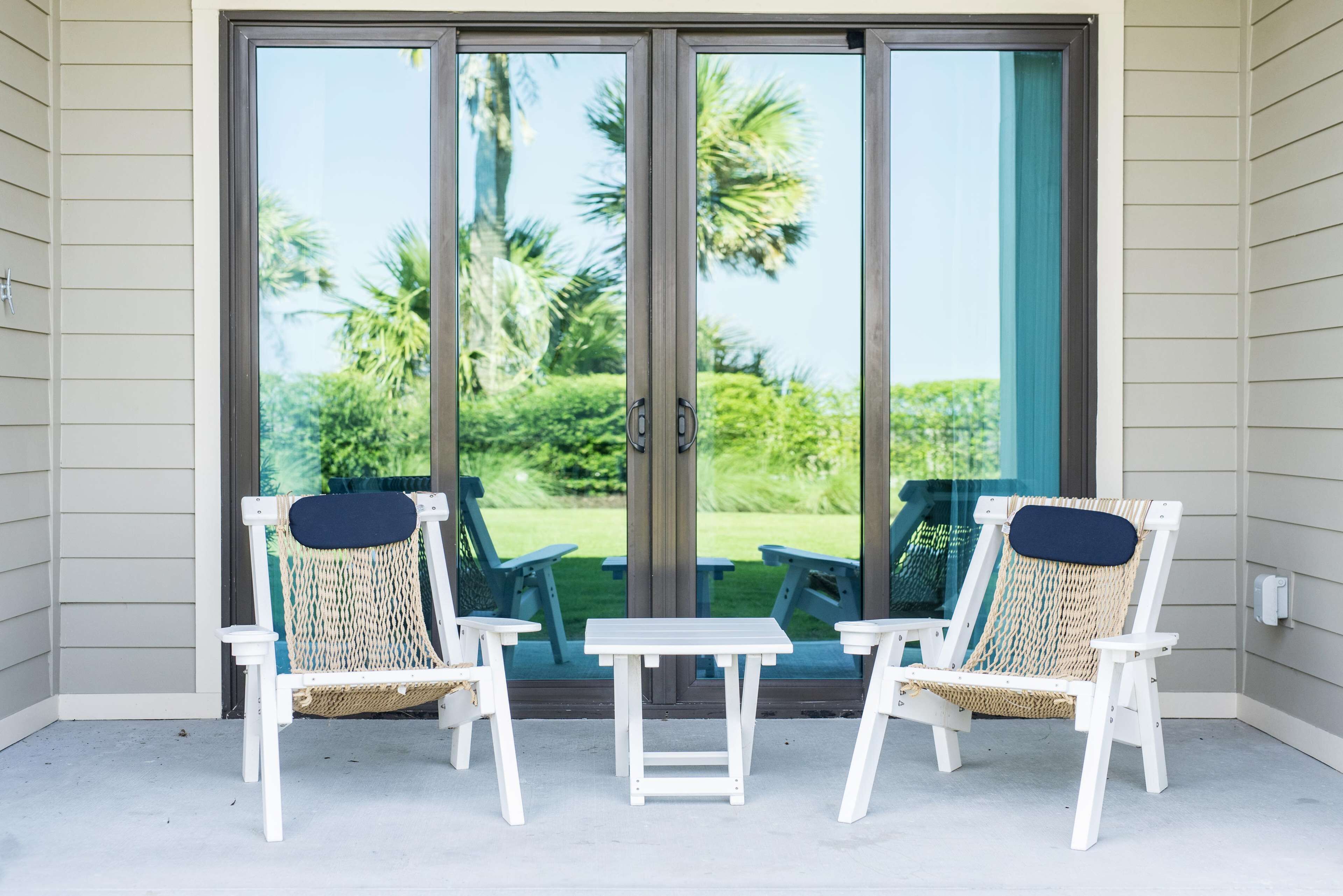 A covered patio with two white chairs and a small table, overlooking a lush green lawn and palm trees through large sliding glass doors.