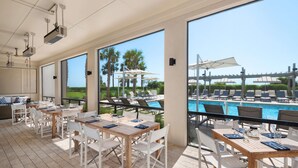 A restaurant with outdoor seating overlooking a pool, featuring large windows and a view of palm trees. - Jekyll Ocean Club (Jekyll Island)