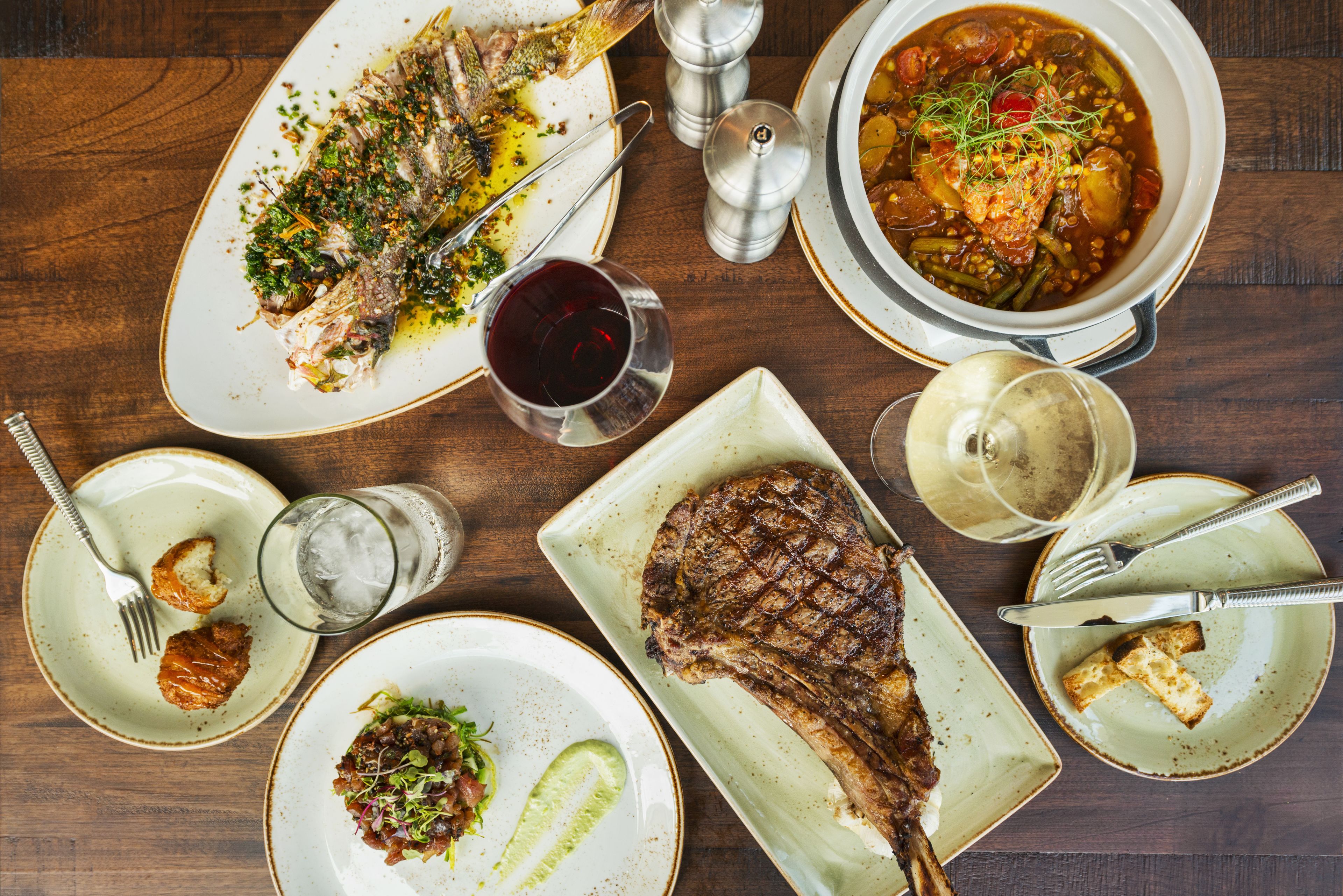 A dining table with various dishes including fish, steak, and a stew, along with cutlery and glasses.