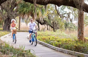Two people riding bicycles on a wooden boardwalk surrounded by greenery and trees.