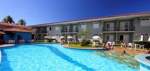 Una piscina con lettini e ombrelloni, circondata da un edificio con numerosi balconi e un tetto in tegole.