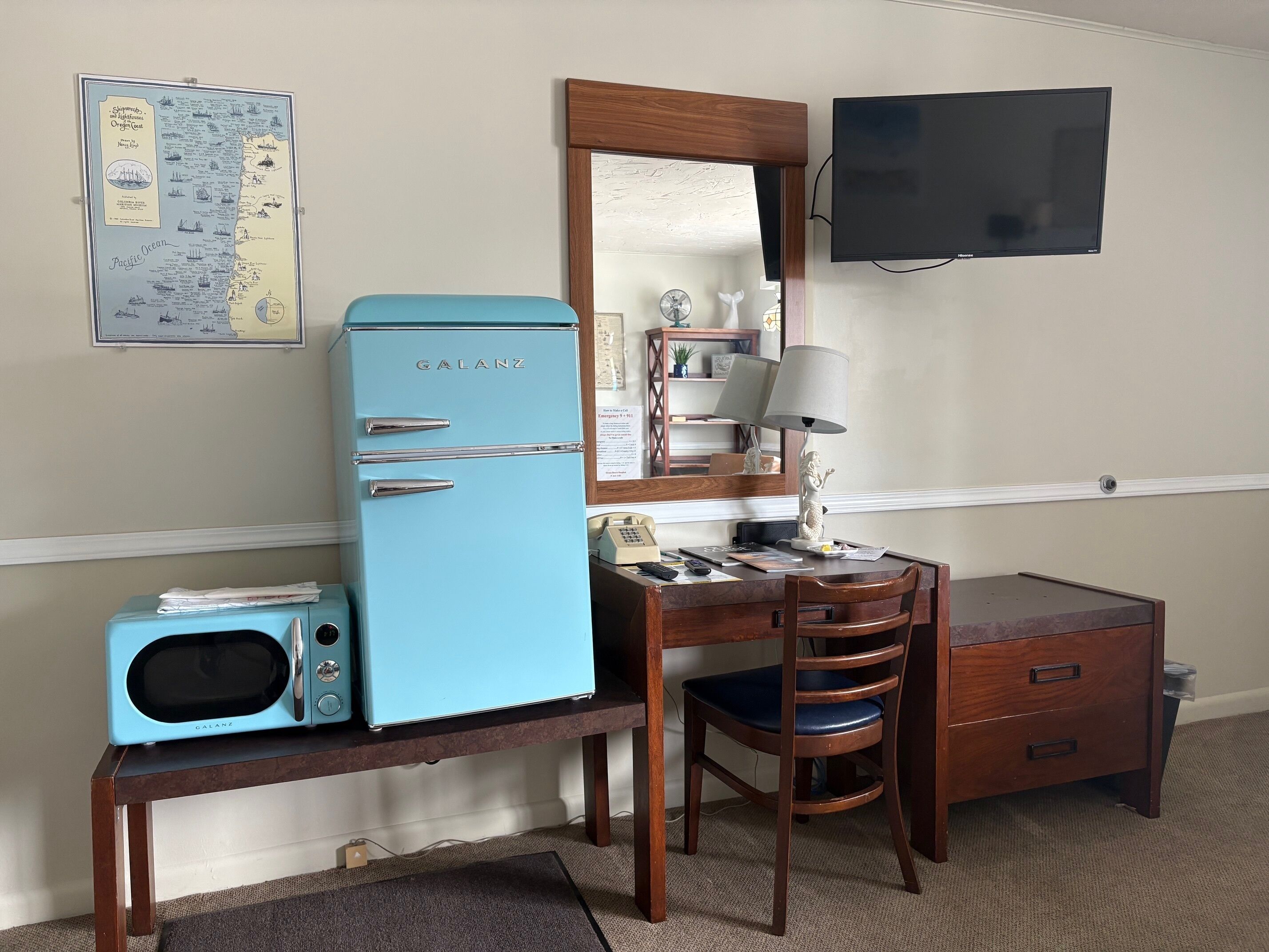 Una habitación con un refrigerador azul antiguo, un escritorio de madera con una silla, un televisor montado en la pared y un mapa enmarcado en la pared.
