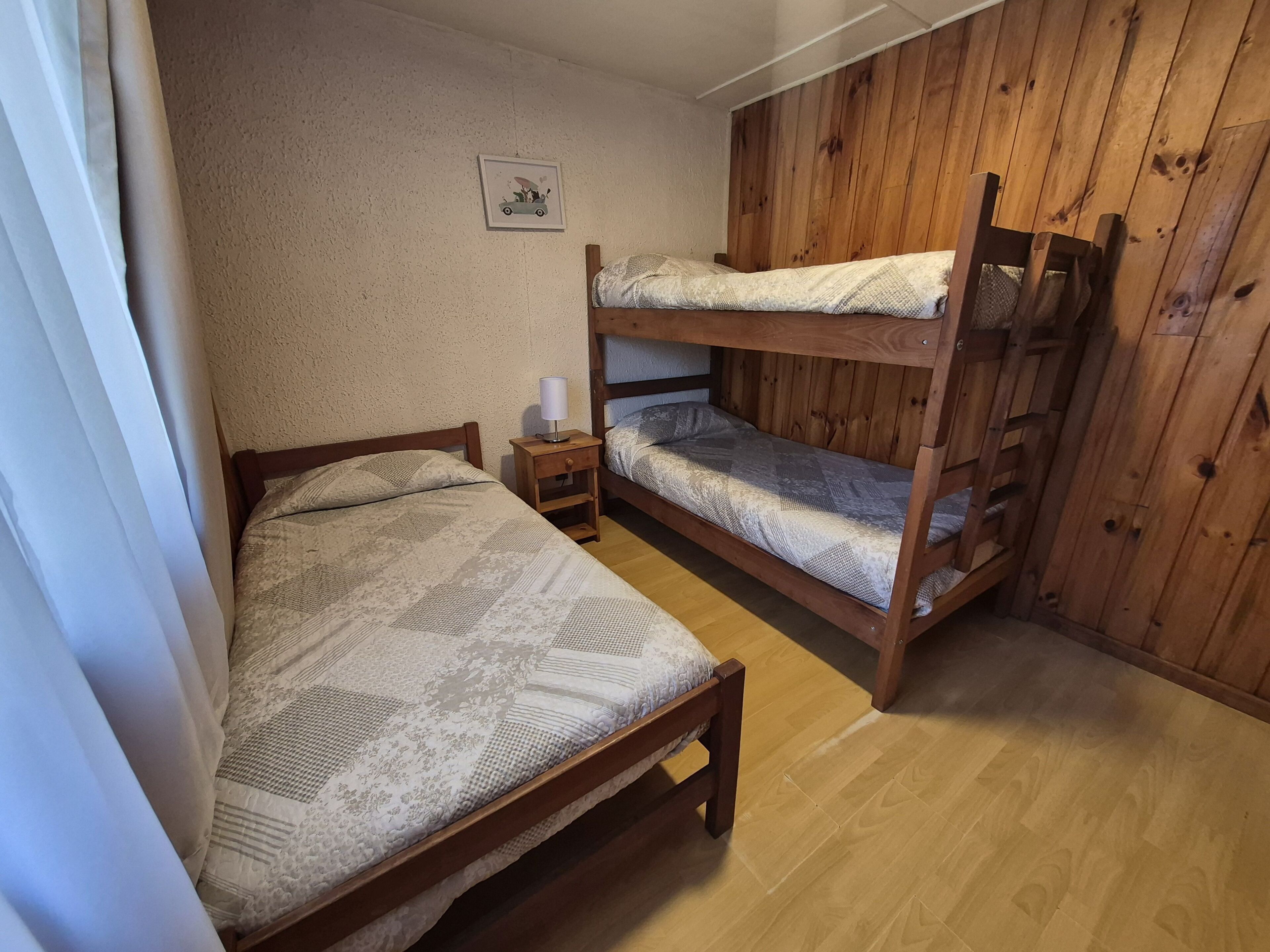 A room with a bunk bed, a single bed, a bedside table with a lamp, and a wall-mounted picture.