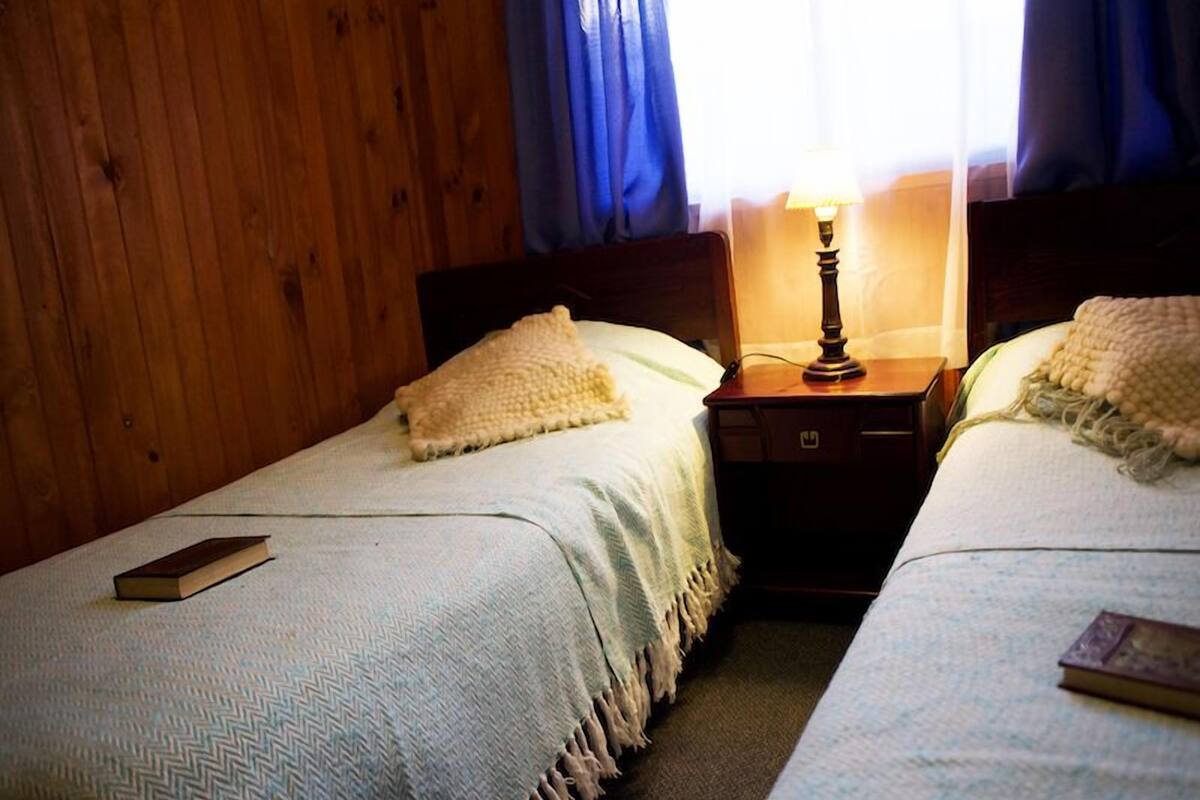 A room with two single beds, wooden walls, a nightstand with a lamp, and a window with curtains.
