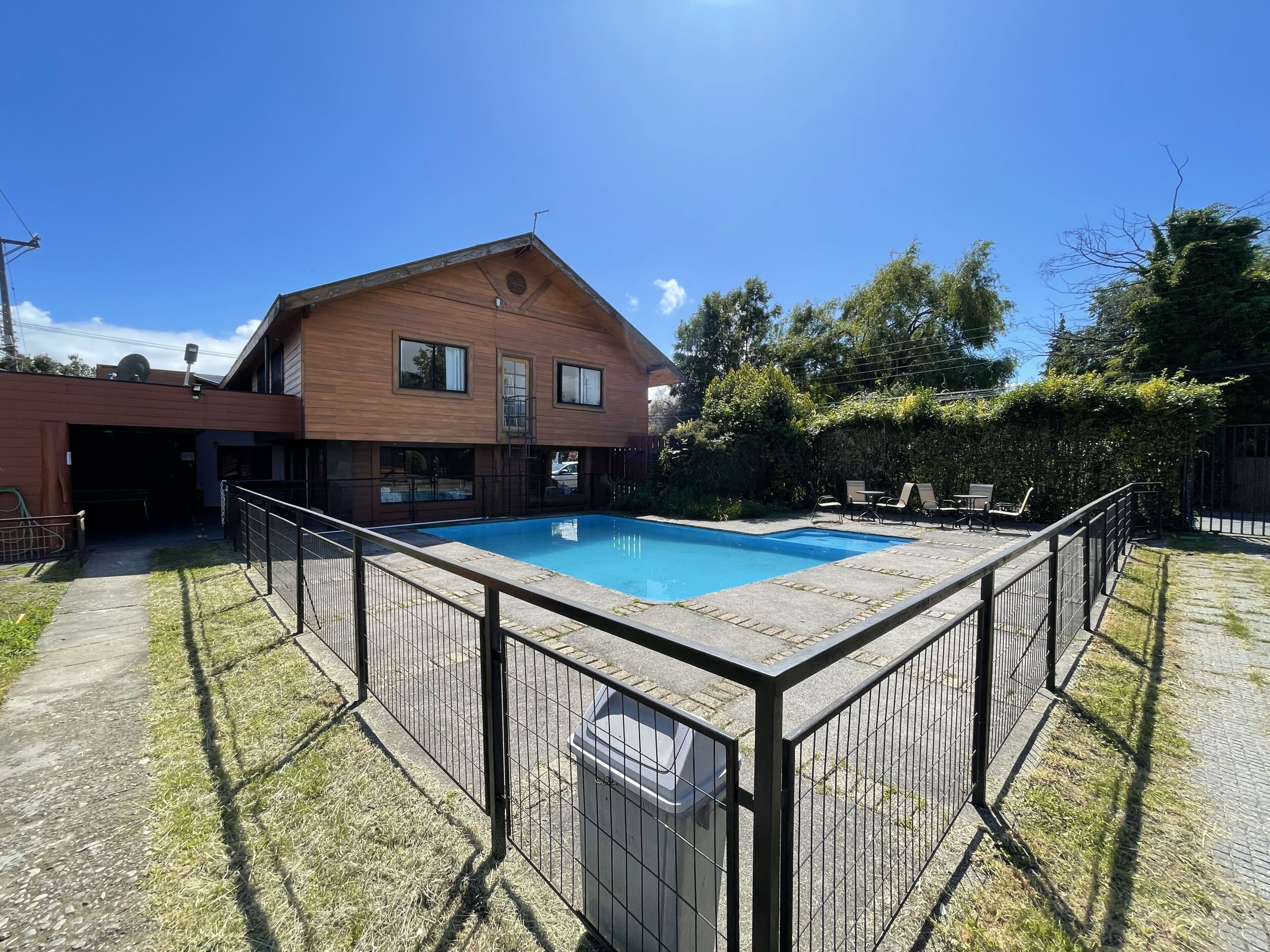 Una piscina rectangular con un deck de madera alrededor, un edificio de ladrillo con ventanas y una zona verde con árboles.