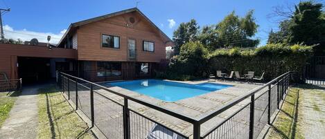 Una piscina rectangular con un deck de madera alrededor, un edificio de ladrillo con ventanas y una zona verde con árboles.