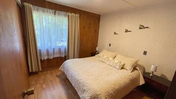 A bedroom with a large bed, wooden flooring, a window with curtains, a wall-mounted light, and a small nightstand.