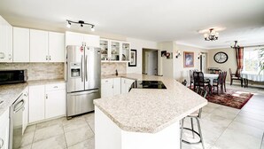 A modern kitchen with white cabinets, stainless steel appliances, a granite countertop, and a dining area with a clock and a window. - Butterfly Manor Bed & Breakfast (Niagara Falls)