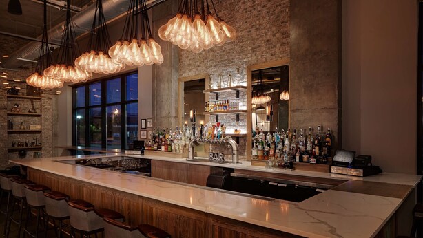 A bar area with a long counter, multiple stools, and a variety of bottles and glasses. - Watt Hotel Rahway, Tapestry Collection by Hilton (Rahway)