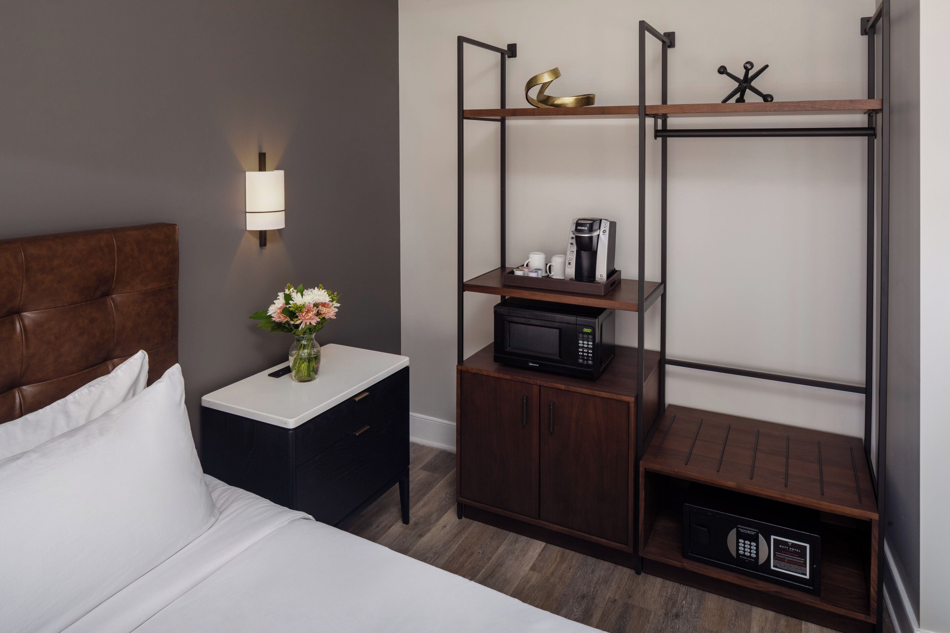 Habitación de hotel con cama, mesita de noche, lámpara y un armario empotrado con cafetera y microondas.