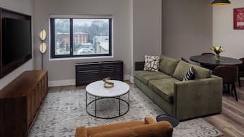 Una sala de estar con sofá, mesa de centro, televisor, mesa de comedor y ventana con vista a edificios.