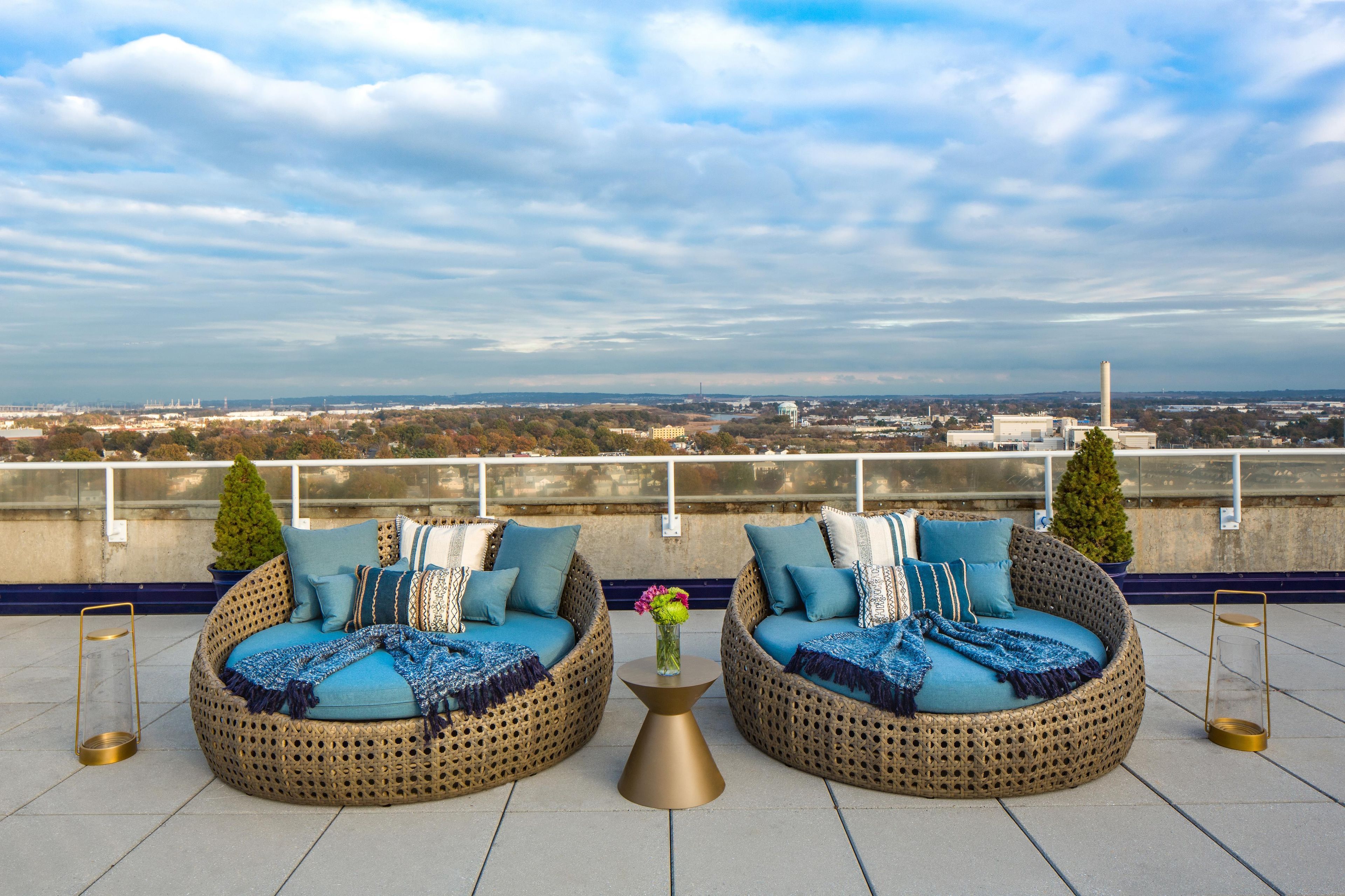 Two wicker sofas with blue cushions and decorative pillows, a small table with a vase of flowers, and a view of a cityscape with buildings and a water tower.