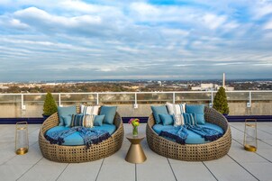 Two wicker sofas with blue cushions and decorative pillows, a small table with a vase of flowers, and a view of a cityscape with buildings and a water tower.