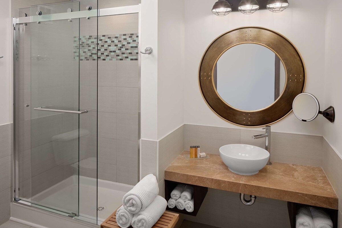 Un baño moderno con cabina de ducha de vidrio, un espejo redondo, un lavamanos blanco y toallas cuidadosamente dobladas.