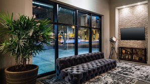 A modern living room with a large window, a potted plant, a tufted sofa, a patterned rug, and a mounted TV. - Watt Hotel Rahway, Tapestry Collection by Hilton (Rahway)