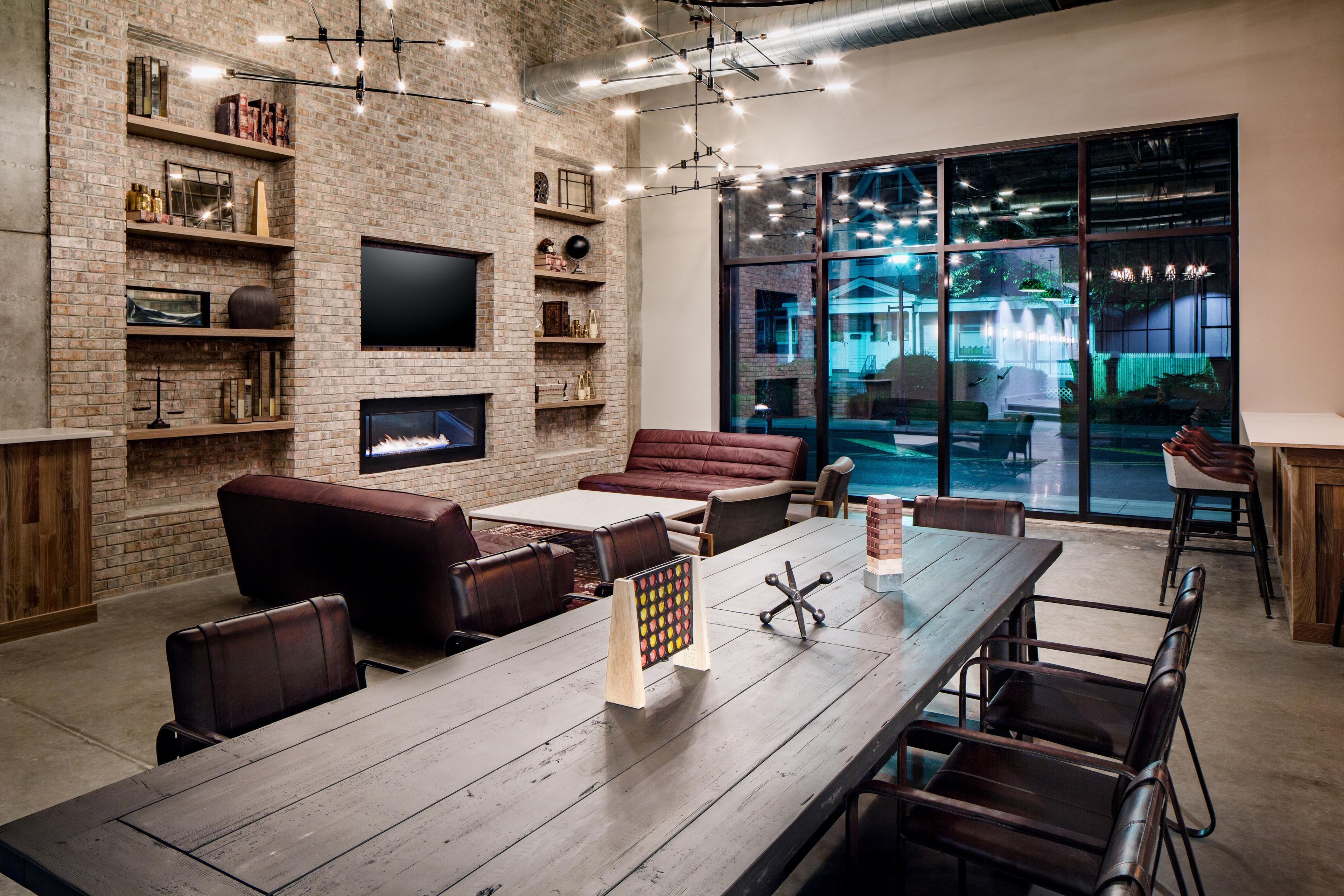 A modern living room with a brick wall, a flat-screen TV, leather sofas, a dining table, and a large window with a view of a house.