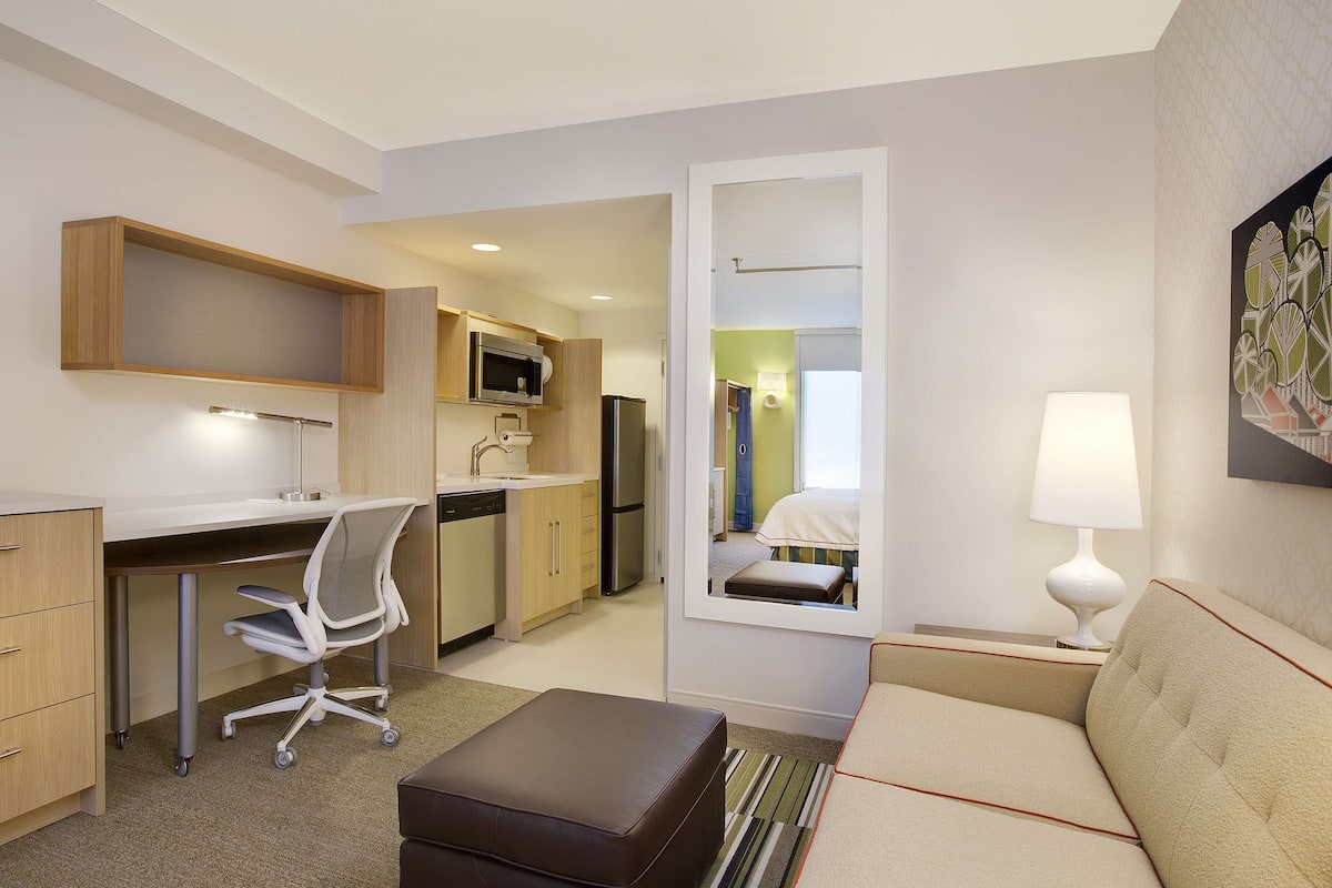 A compact hotel room with a kitchenette, a desk, a chair, a sofa, and a bedside table with a lamp.