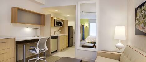 Una habitación de hotel compacta con kitchenette, un escritorio, una silla, un sofá y una mesita de noche con lámpara.
