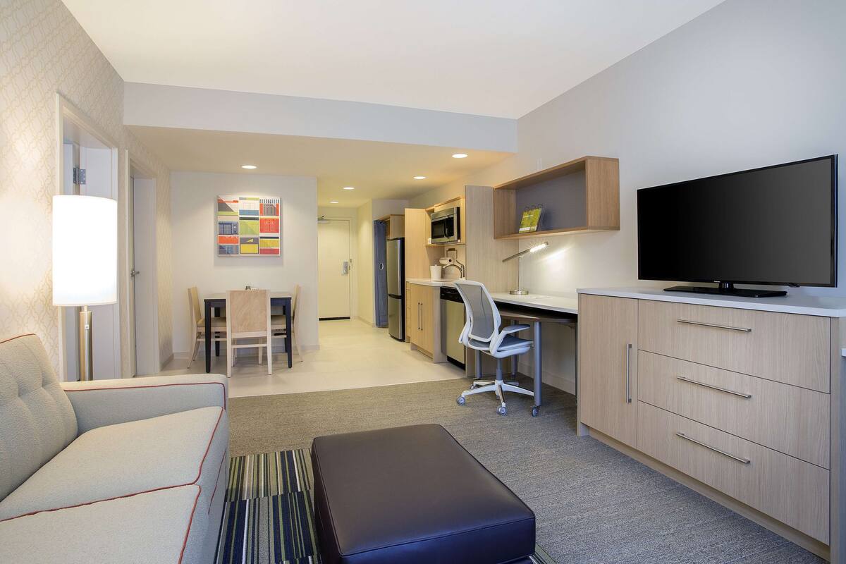 A modern hotel room with a sofa, a desk, a television, and a kitchenette.