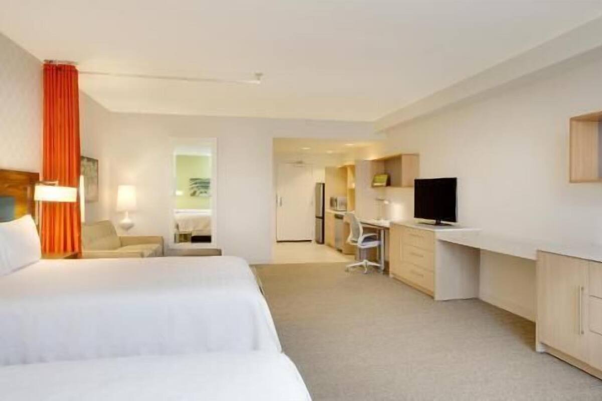 A hotel room with two beds, a desk, a television, and a kitchenette.