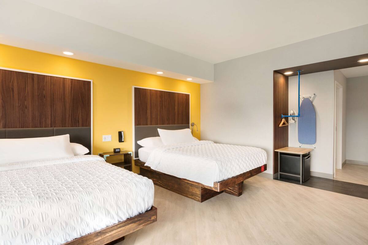 A modern hotel room with two beds, wooden headboards, and a small bedside table with a lamp.