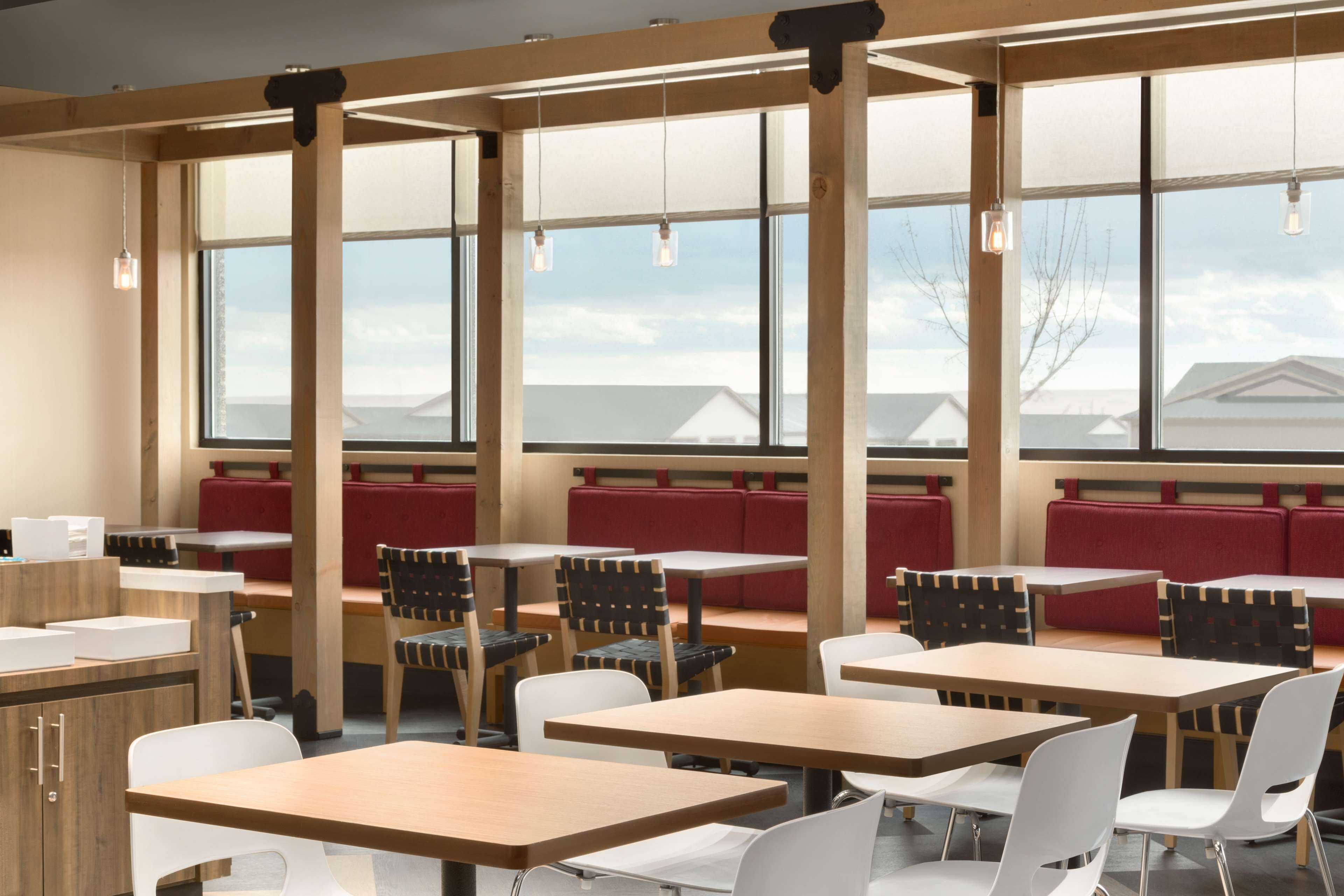 Un restaurante con mesas de madera, asientos tapizados en rojo, ventanas amplias y lámparas colgantes.