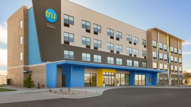 A modern hotel building with a prominent 'tru' logo and 'Hilton' branding. It features a flat roof, multiple windows, and a blue and beige color scheme.