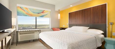 Une chambre d’hôtel moderne dotée d’un grand lit, d’un bureau avec un téléviseur à écran plat, d’une fenêtre donnant sur la ville et d’un tableau abstrait aux couleurs vives accroché au mur.