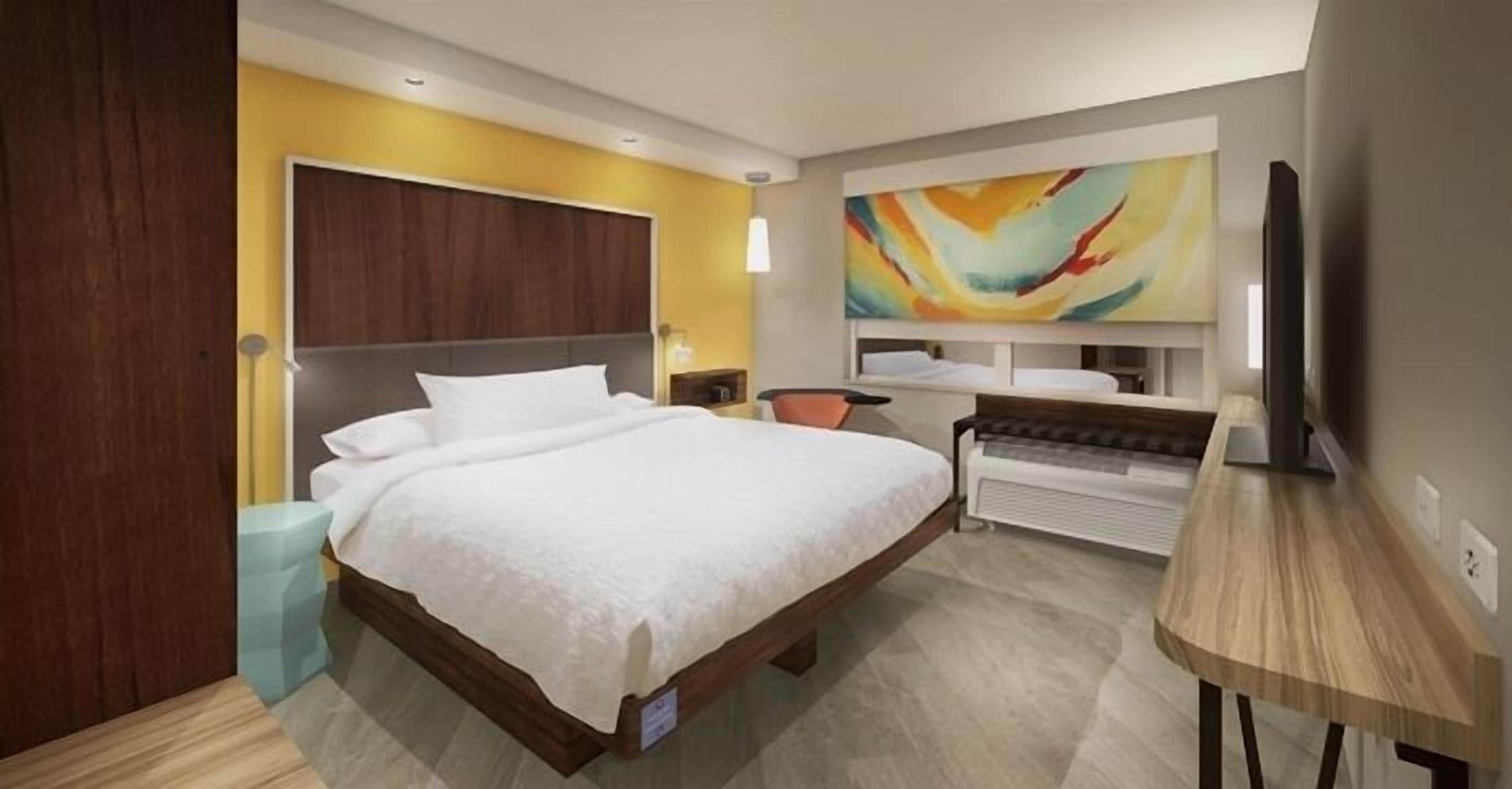 A modern hotel room with a large bed, a desk, a television, and a colorful abstract painting on the wall.
