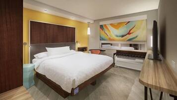 A modern hotel room with a large bed, a desk, a television, and a colorful abstract painting on the wall.