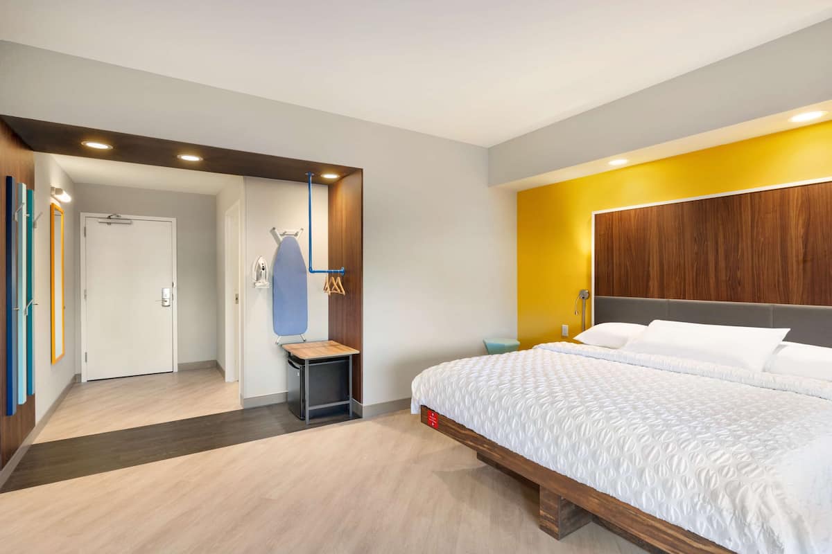 A modern hotel room with a large bed, a wooden headboard, a small side table, and a wardrobe.