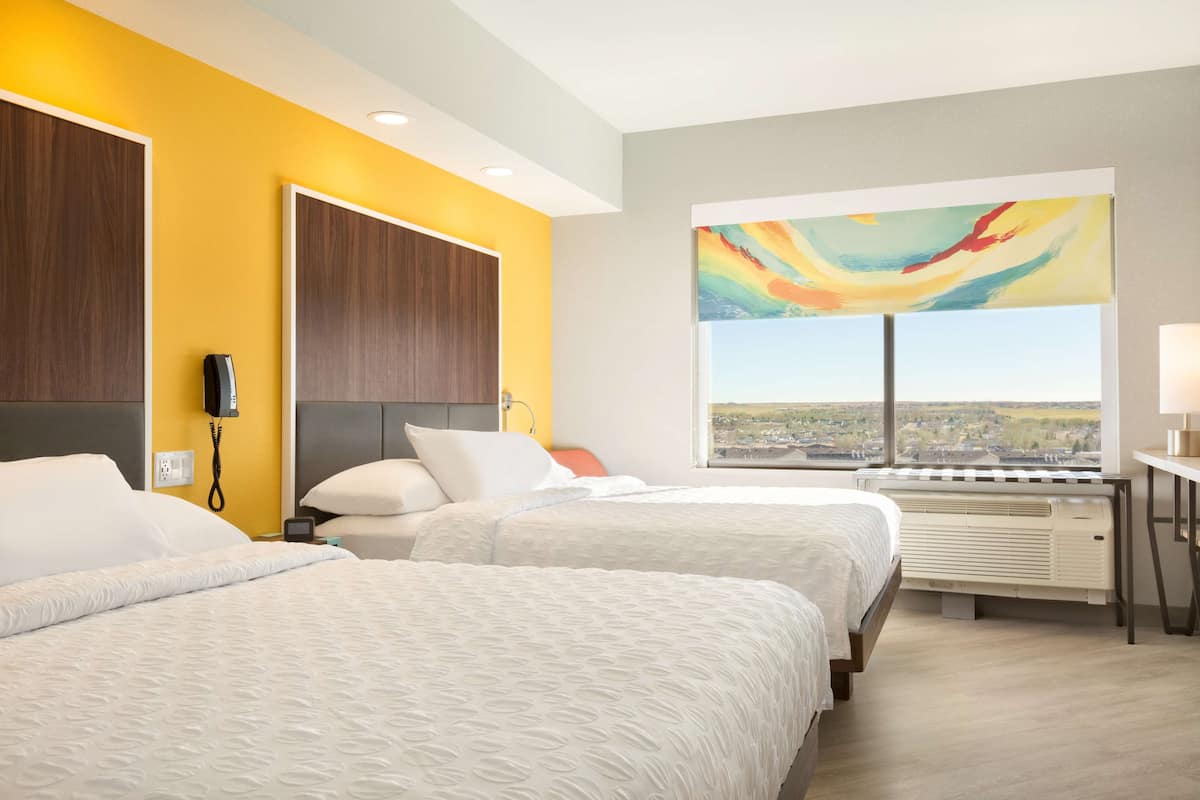 A hotel room with two beds, a large window with a city view, a painting on the wall, and a desk with a lamp.