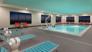 A modern indoor pool area with multiple lounge chairs and tables, large windows with night views, and a ceiling with recessed lighting.
