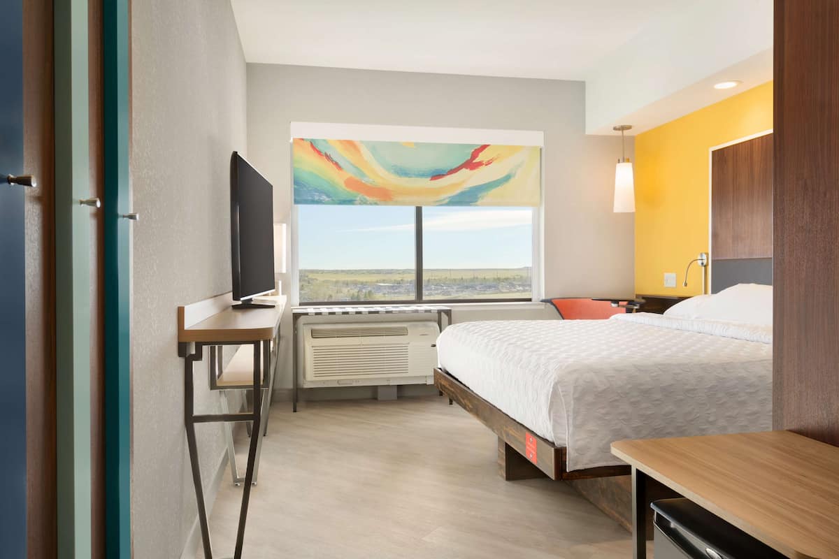 A hotel room with a bed, a desk with a TV, a window with a cityscape view, and a painting on the wall.