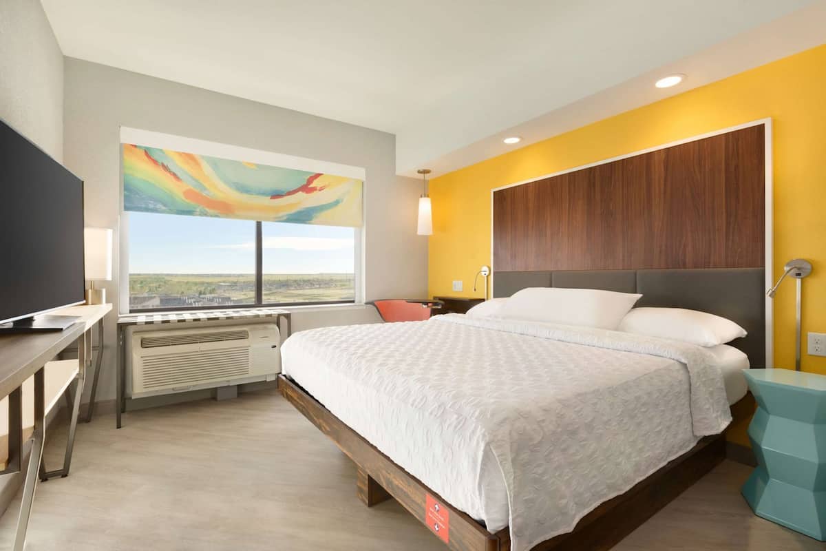 A hotel room with a bed, a desk, a TV, a window with a view, and a painting on the wall.