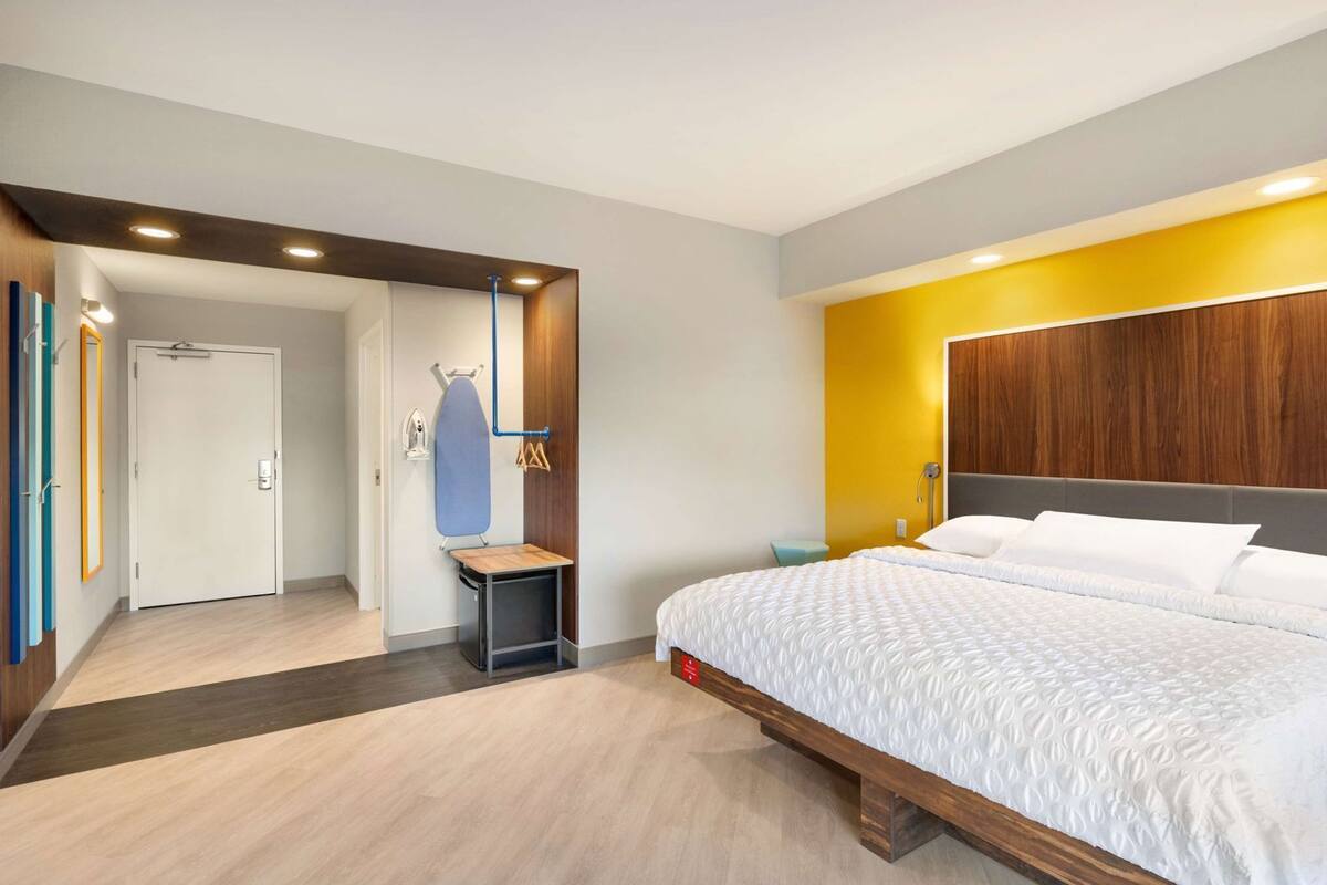 A modern hotel room with a bed, a wooden headboard, a small table with an ironing board, and a door leading to another room.