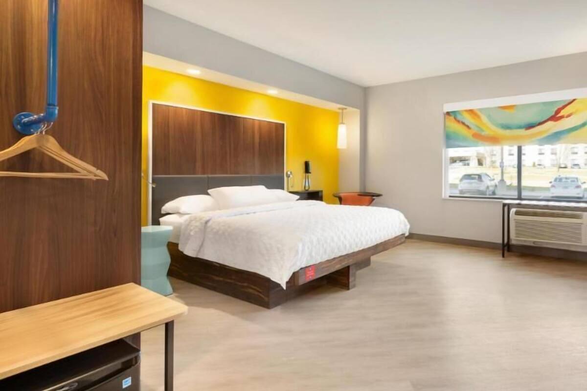 A modern hotel room with a bed, a wooden dresser, a hanging coat rack, a bedside table, and a window with a view of the street.