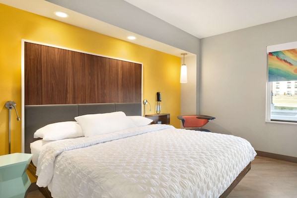 A modern hotel room with a large bed, a wooden headboard, a bedside table, a chair, and a window with a view.