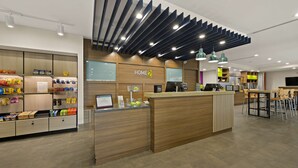 A modern hotel lobby with a wooden reception desk, a self-service snack station, and a seating area with tables and chairs. - Home2 Suites by Hilton Baytown (Baytown)