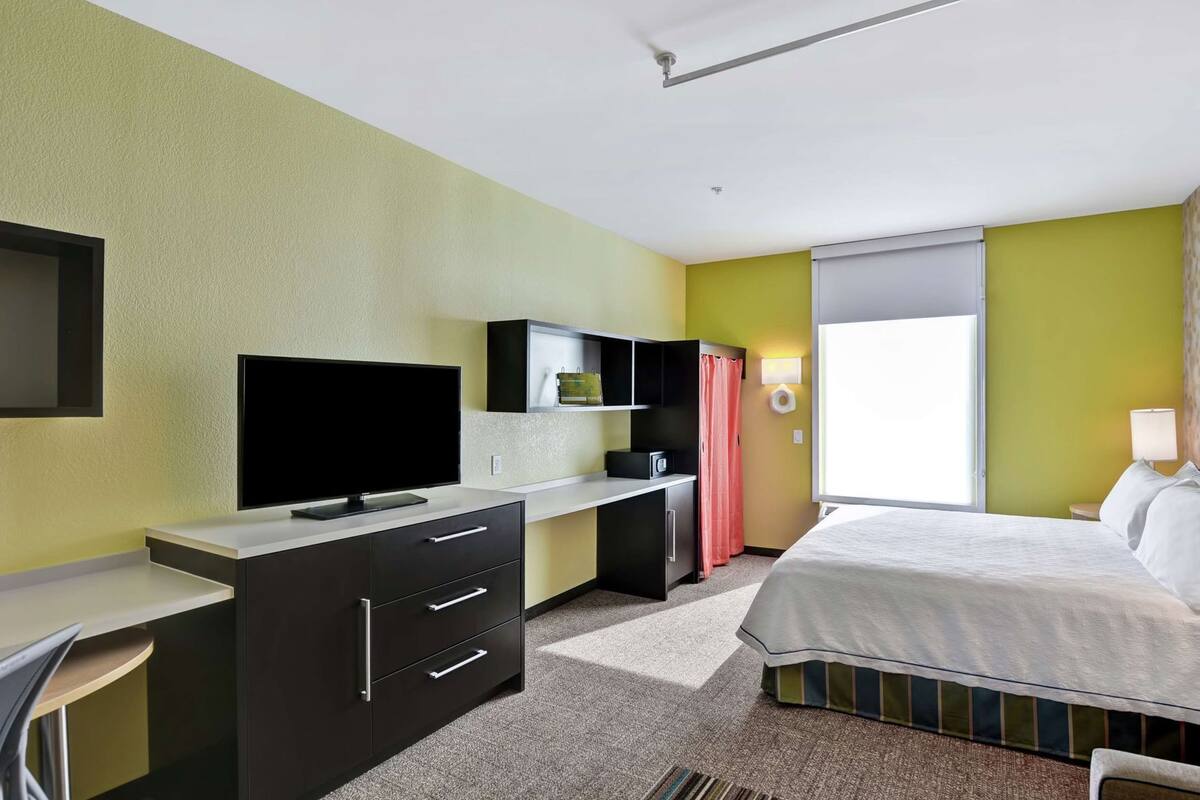 Een hotelkamer met een bed, een bureau met een televisie, een klein kastje en een raam met gordijnen.