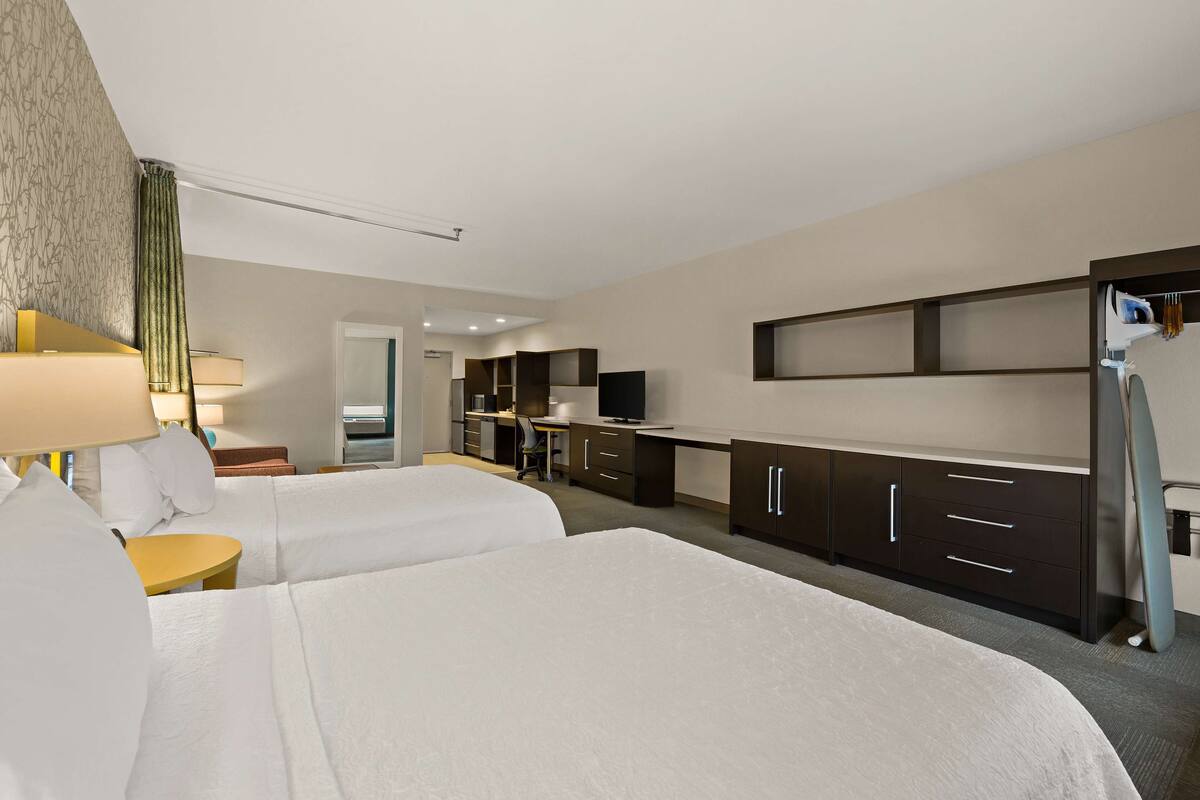 A hotel room with two beds, a desk, a chair, a TV, and a kitchenette.