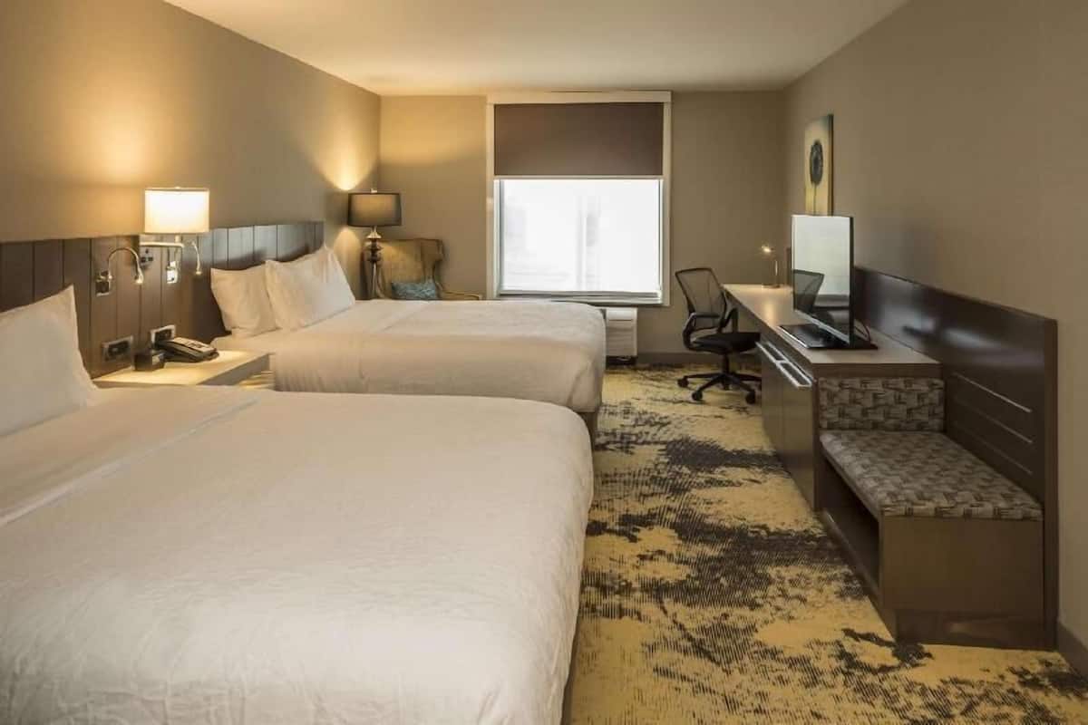 A hotel room with two beds, a desk, a chair, a TV, and a window.