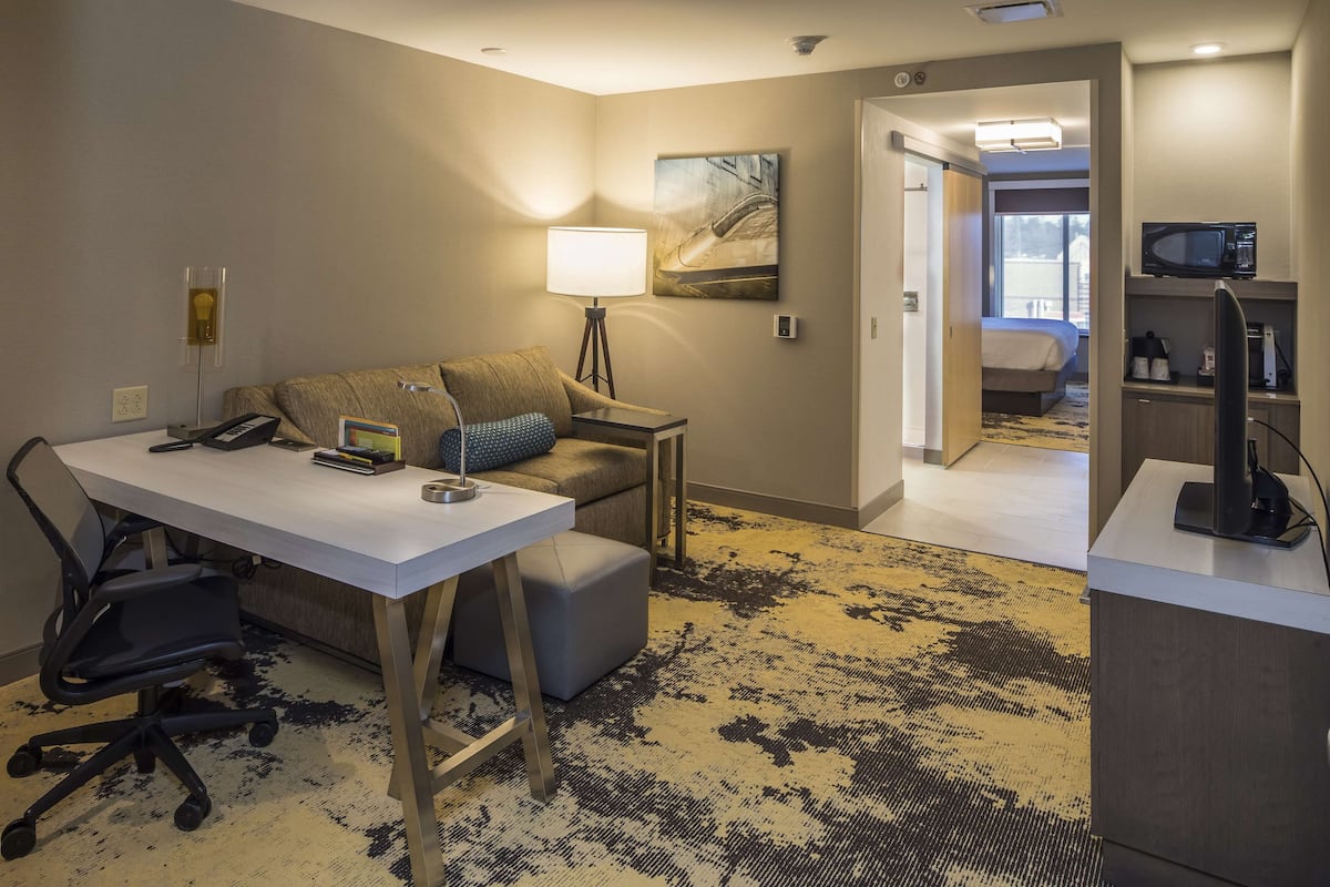 A hotel room with a desk, chair, lamp, and a bed in the background.