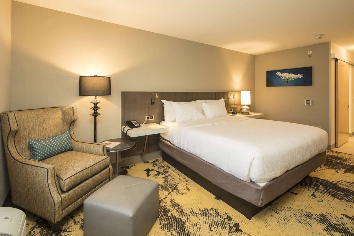 A hotel room with a large bed, a chair, a small table, and a lamp.
