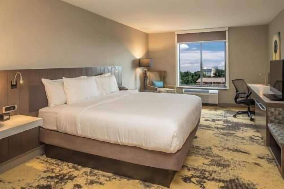 A hotel room with a large bed, a desk, a chair, and a window with a city view.