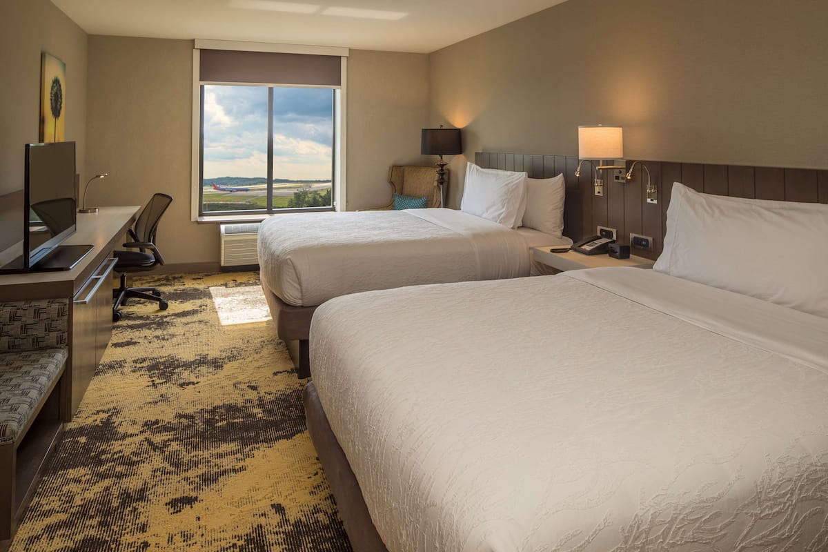 A hotel room with two beds, a desk with a computer, a chair, a lamp, and a window with a view of the airport.
