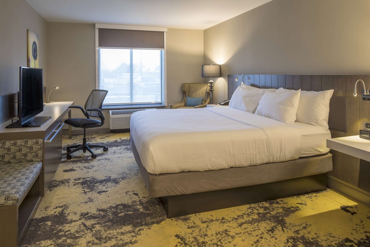 A hotel room with a large bed, a desk with a computer, a chair, a TV, and a window with a view.