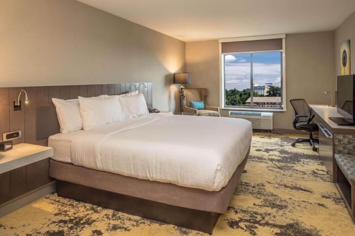 A hotel room with a large bed, a desk, a chair, a lamp, and a window with a city view.